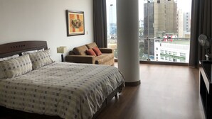 A hotel room with a large bed, a sofa, a window with a city view, and a fan. - Kennedy Park Miraflores apartment (Miraflores)