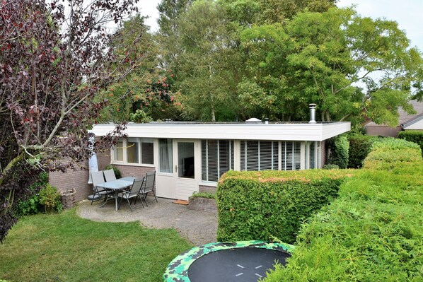 Une petite maison de plain-pied, à l’extérieur blanc, dotée d’un patio couvert avec une table et des chaises, et d’un jardin bien entretenu agrémenté d’une haie et d’un trampoline.