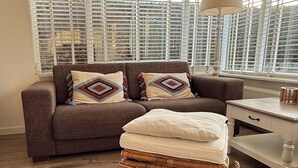 A living room with a brown sofa, patterned cushions, a wicker ottoman, a white side table, and a window with blinds. - Cozy bungalow in rural area near beautiful dunes, beach and sea. (Schoorldam)