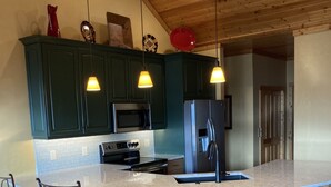 Una cocina con armarios verde oscuro, una isla grande con un lavamanos, un refrigerador y un microondas. Hay dos lámparas colgantes sobre la isla.