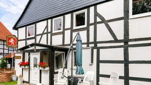 A traditional half-timbered building with a white facade and black beams. There are three white plastic chairs arranged outside, a blue umbrella, and a wooden fence. - Historic coach house is looking forward to two and four-legged friends (Coppenbrügge)