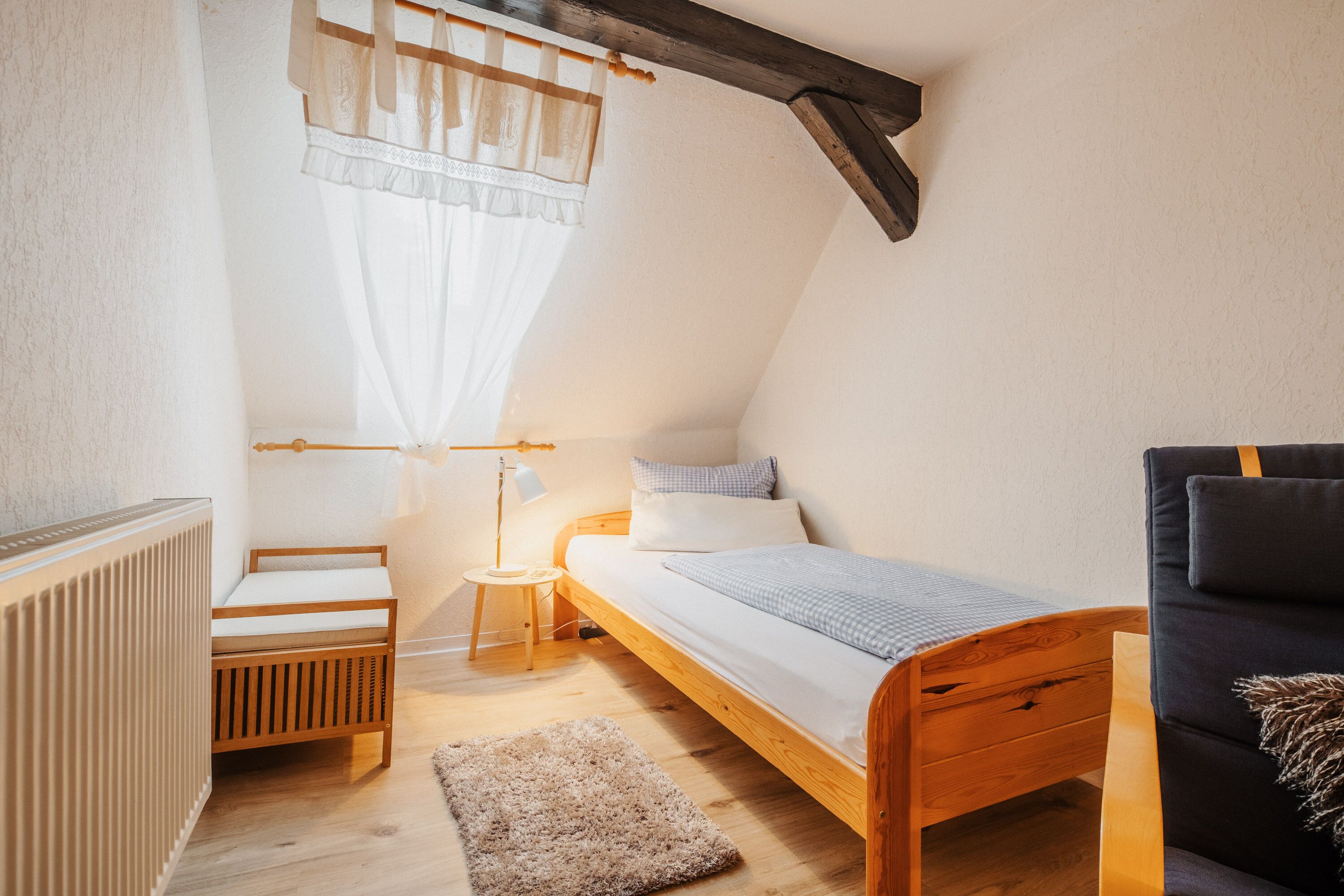 Un dormitorio pequeño y acogedor con techo inclinado, una cama individual, una silla de madera, una mesita y un radiador.