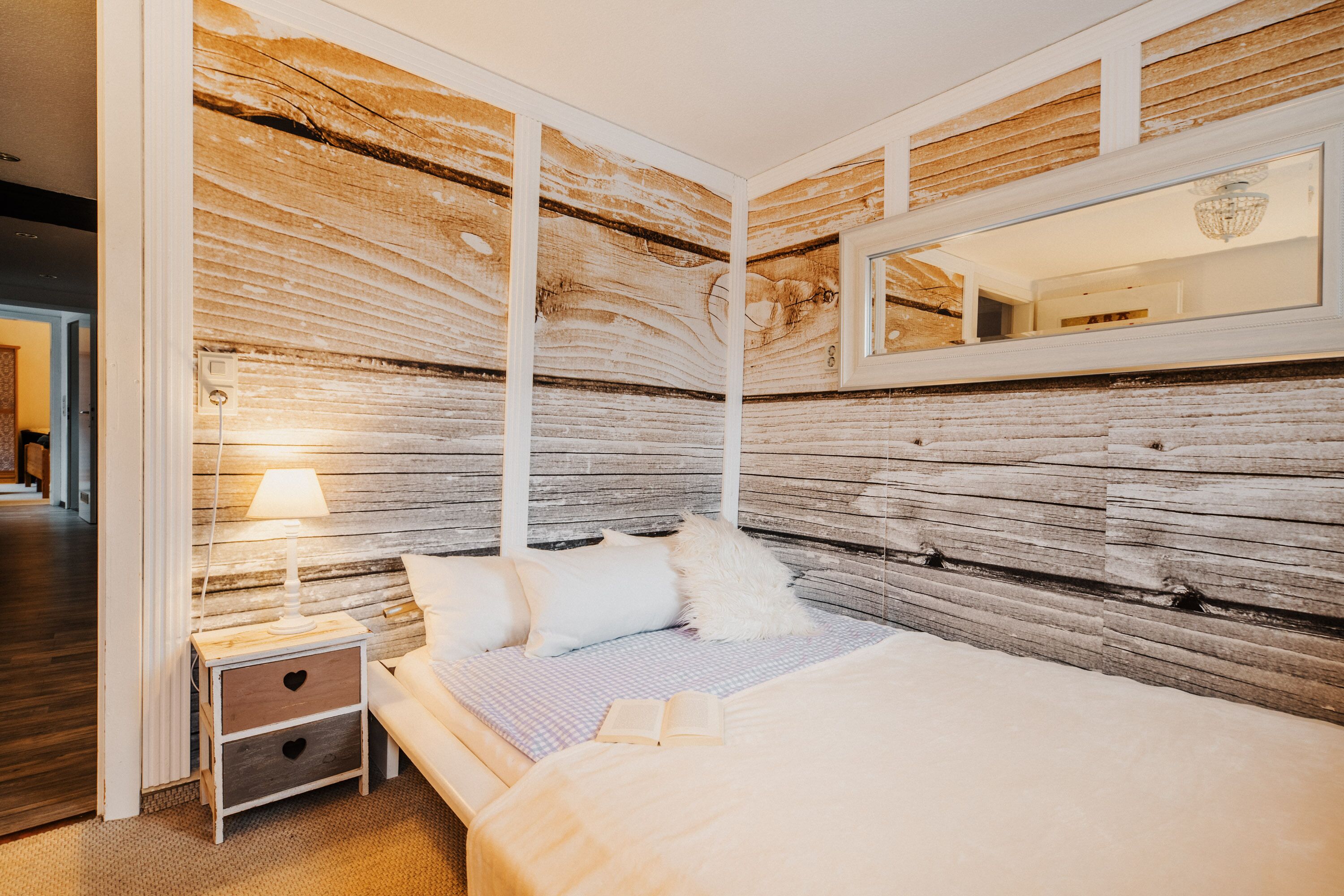 A bedroom with a wooden wall panel, a bed with pillows, a bedside table with a lamp, and a mirror above the bed.