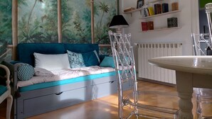 Una habitación con un sofá azul, una silla transparente, un radiador y una estantería con libros y marcos con fotografías.