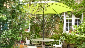 Un patio con una mesa y sillas, rodeado de abundante vegetación y un gran paraguas verde.