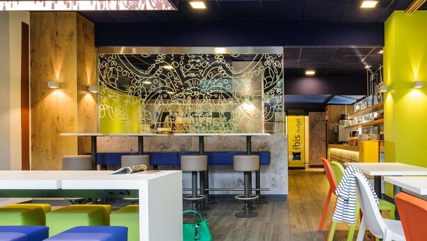 A modern cafe with colorful seating, a bar area, and a decorative wall with abstract patterns. - Ibis Budget Muenchen Airport Erding (Erding)
