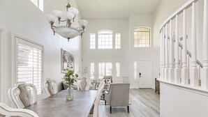 Ein großzügiger Essbereich mit einem langen Holztisch, weißen Polsterstühlen und einem Kronleuchter. Große Fenster und eine Treppe mit weißen Geländern sind ebenfalls vorhanden.