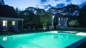A swimming pool with lounge chairs and a covered area, surrounded by trees and a house with illuminated windows. - East Hampton Luxury: Heated Pool, Hot Tub and Expansive Yard with 6 Bedrooms (East Hampton)