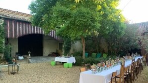 Un espacio de comedor al aire libre con una larga mesa dispuesta para una comida, rodeado de árboles y un edificio con techo de tejas.