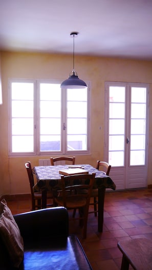 Un espace repas comprenant une table et des chaises, un luminaire suspendu et deux fenêtres à cadre blanc.