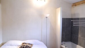 Ein Schlafzimmer mit einem Bett, einer Nachttischlampe, einem kleinen hölzernen Hocker und einem Badezimmer mit einer Badewanne und einem Handtuchhalter.
