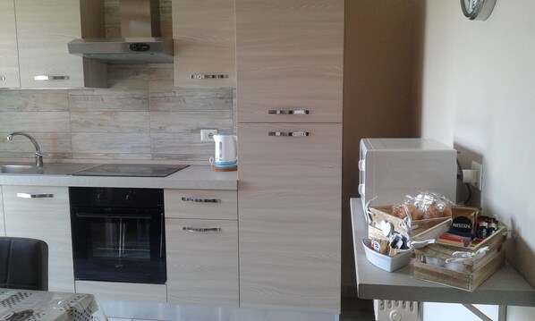 A modern kitchen with a built-in oven, a kettle, and a coffee machine. There is a wooden cabinet with a handle and a small wooden table with a basket containing various items.