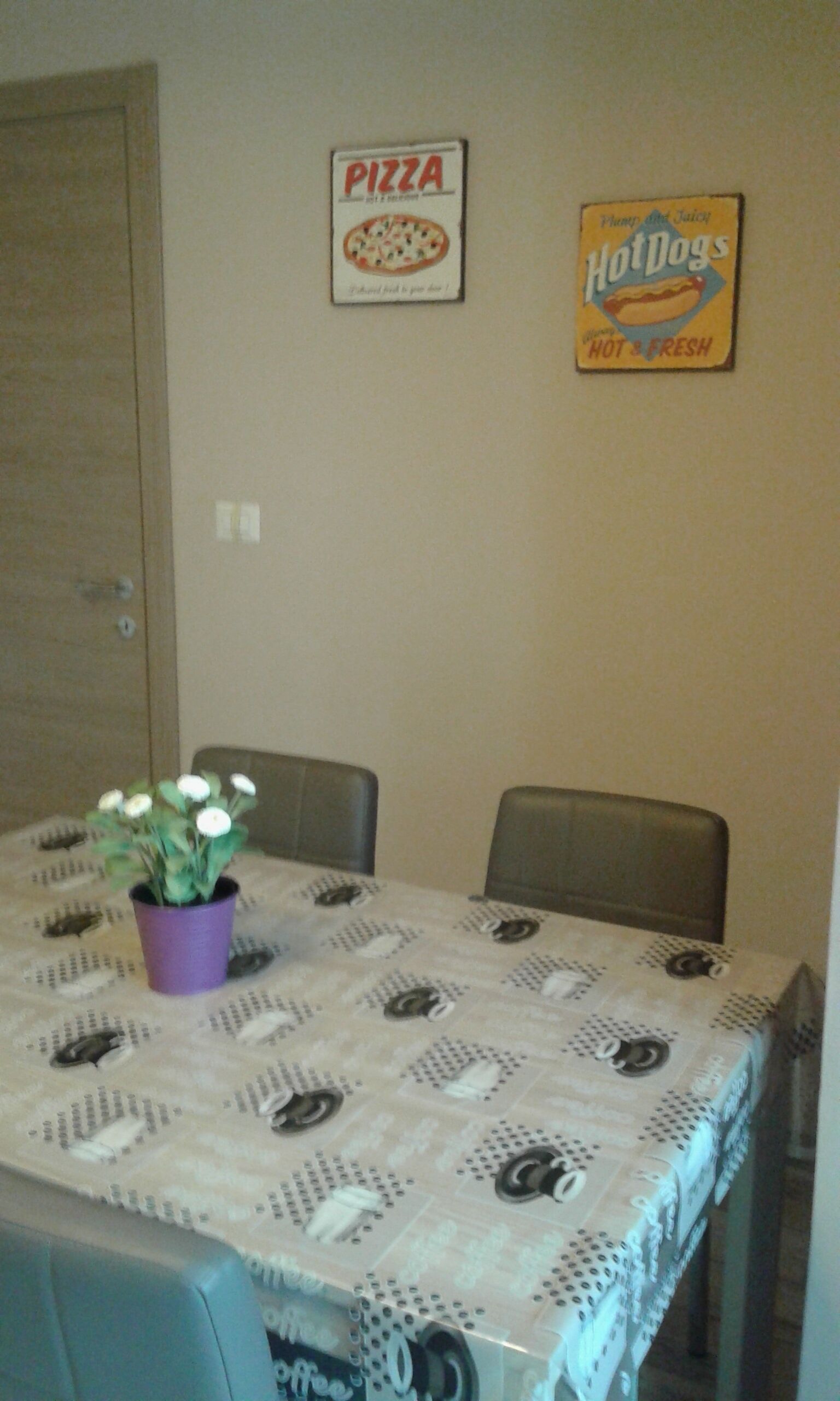 Un coin repas avec une table recouverte d’une nappe à motifs, deux chaises, une plante en pot et deux panneaux muraux faisant la publicité de pizzas et de hot-dogs.