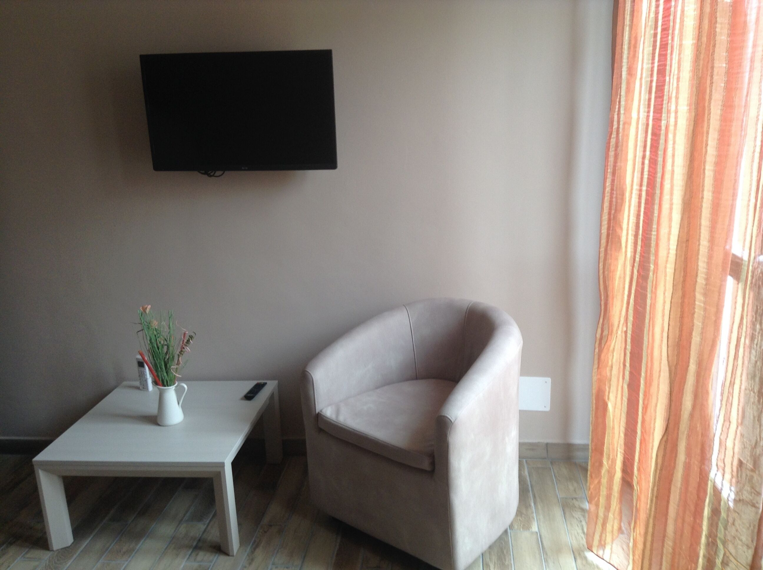 A living room with a flat-screen TV mounted on the wall, a small white table with a vase and remote, and a beige armchair.