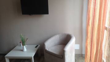 A living room with a flat-screen TV mounted on the wall, a small white table with a vase and remote, and a beige armchair.