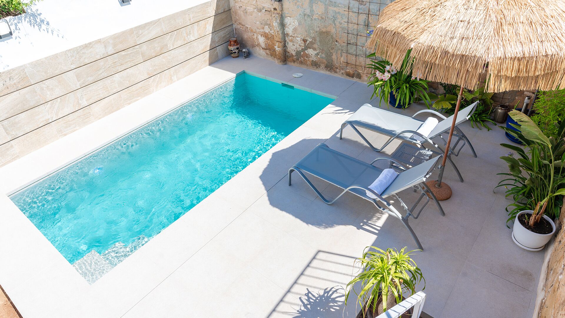 A rectangular swimming pool with clear blue water, surrounded by a tiled deck. There are two lounge chairs with cushions, a thatched umbrella, and several potted plants.