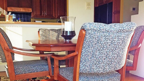 A dining area with a wooden table and two patterned chairs, a glass candle holder, and a kitchen in the background.
