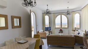 A living room with a dining table, chairs, a sofa, and a television. There are large windows with curtains, a chandelier, and a radiator. - Acropolis Sea View Villa - Panoramic Views & Stunning location (Poli Crysochous)