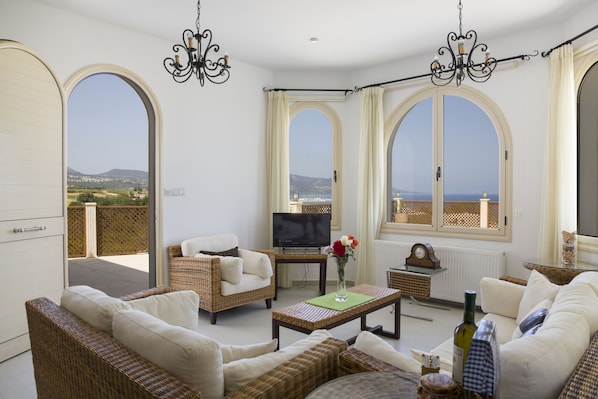 A spacious living room with a television, wicker furniture, a coffee table, and large windows offering a view of the sea. - Acropolis Sea View Villa - Panoramic Views & Stunning location (Poli Crysochous)