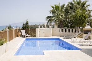 A rectangular swimming pool with a clear blue water surface, surrounded by a tiled deck. There are several lounge chairs arranged around the pool, and a wooden fence with decorative elements. In the background, there are palm trees and a view of the sea and distant mountains. - Acropolis Sea View Villa - Panoramic Views & Stunning location (Poli Crysochous)