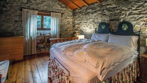 A rustic bedroom with a stone wall, wooden floor, a bed with floral bedding, a nightstand, and a window with curtains.