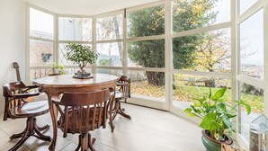 Un comedor con una mesa redonda y sillas, ventanas amplias con vistas a árboles y un edificio, y una planta en maceta en el alféizar.