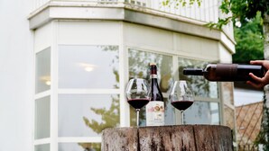 Una botella de vino siendo servida en dos copas sobre un tocón de madera, con un edificio al fondo.