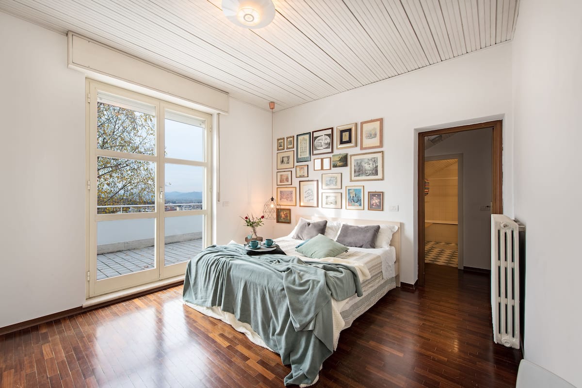 A bedroom with a large window, a bed with a green blanket, and a wall with framed pictures.