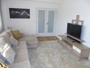 A living room with a beige sofa, a television on a wooden stand, and a door with frosted glass panels. - Oporto Beach Apartment (Duplex) (Gulpilhares)