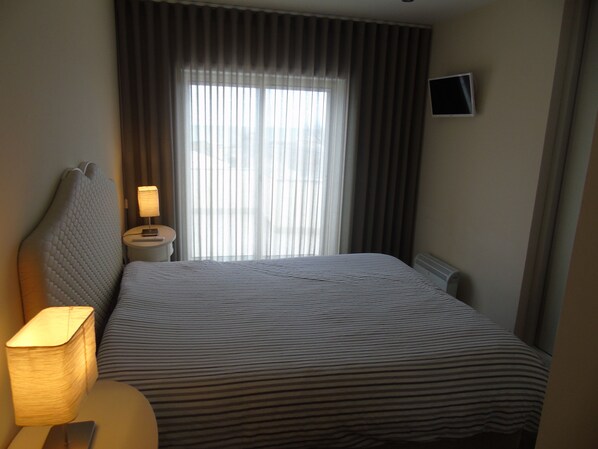 Ein Hotelzimmer mit einem Bett, einem Nachttisch mit Lampe, einem an der Wand montierten Fernseher und einem Fenster mit Vorhängen.