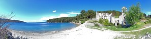 A coastal landscape with a clear blue sea, a rocky beach, and ancient ruins with a path leading to them.
