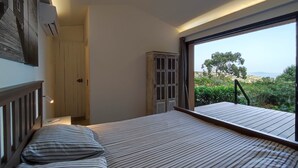 Un dormitorio con un ventanal grande, una cama con ropa de cama a rayas, una puerta de madera y vistas a zonas verdes y colinas.