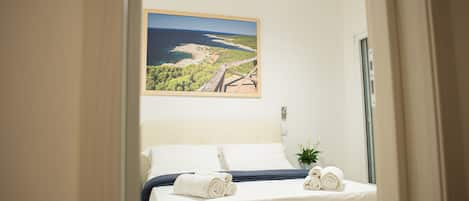 Una cama bien tendida con toallas enrolladas, un cuadro de paisaje enmarcado encima y una lámpara en forma de estrella.