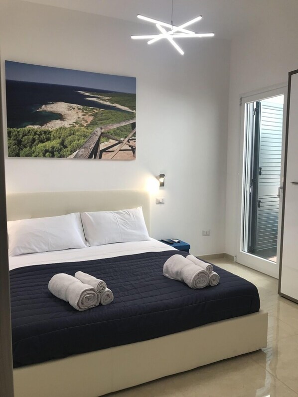A modern bedroom with a large bed, a painting of a coastal landscape, a star-shaped ceiling light, and a view through a glass door.