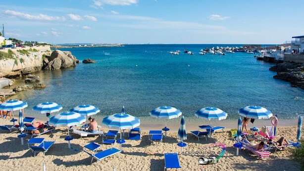 Une plage avec des parasols à rayures bleues et blanches, des chaises longues et une côte rocheuse.