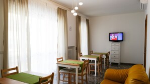 A dining area with wooden tables and chairs, a television on a stand, and a yellow sofa. - Large apartment in the center of Lecce (Lecce)