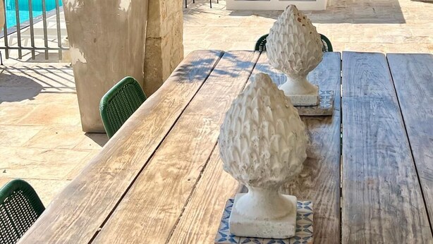 Un patio con una mesa de piedra, dos jarrones decorativos, una parrilla y una piscina al fondo.