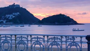 A coastal view with a railing in the foreground, a sunset over hills, and boats on the water. - BartoloBeachApartment. 100m from La KONTXA Beach (Donostia)
