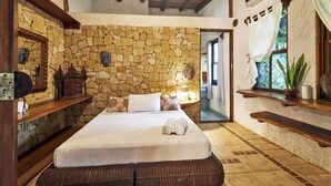 Un dormitorio con pared de piedra, una cama, una mesita de noche, un espejo, una ventana con cortinas y una estantería con una planta.