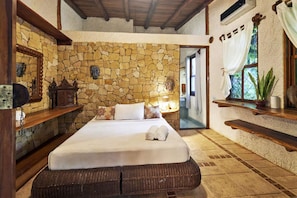 A bedroom with a stone wall, a bed, a nightstand, a mirror, a window with curtains, and a shelf with a plant.