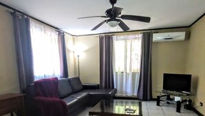 A living room with a sofa, a coffee table, a TV, a ceiling fan, and an air conditioning unit.