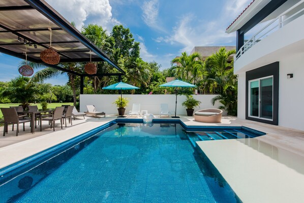A swimming pool with lounge chairs and umbrellas, surrounded by greenery and a modern building.