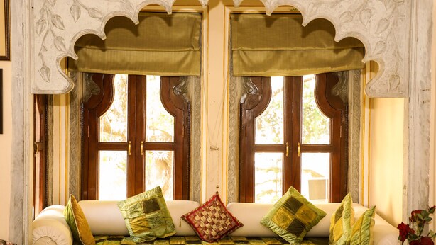 A traditional room with a sofa bed, ornate window frames, and a decorative pillow. - Venus Heritage in the family  (Udaipur)