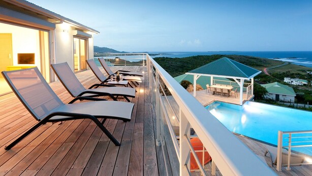 A wooden deck with lounge chairs, a pool, and a view of the ocean. - Les Alizés, magnificent villa with breathtaking view (Oyster pond)