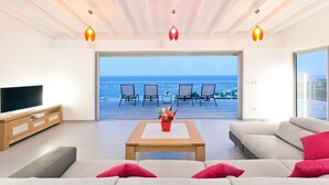 A modern living room with a large TV, a sofa, a coffee table, and a view of the ocean through a glass door. - Les Alizés, magnificent villa with breathtaking view (Oyster pond)