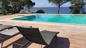 A pool with a view of the sea, surrounded by lounge chairs and trees.