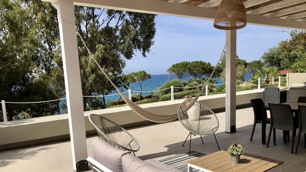 A covered outdoor seating area with a view of the sea, a hammock, and a table with chairs.
