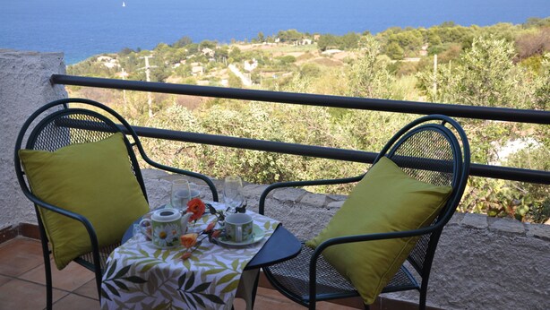 Un balcon avec deux chaises et une table dressée pour deux personnes, offrant une vue sur un paysage côtier avec la mer et la végétation.
