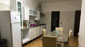 Eine kompakte Küche mit weißen Schränken, einem Kühlschrank, einer Mikrowelle und einem Spülbecken. Ein Esstisch mit vier Stühlen und einer blauen Schüssel steht daneben.