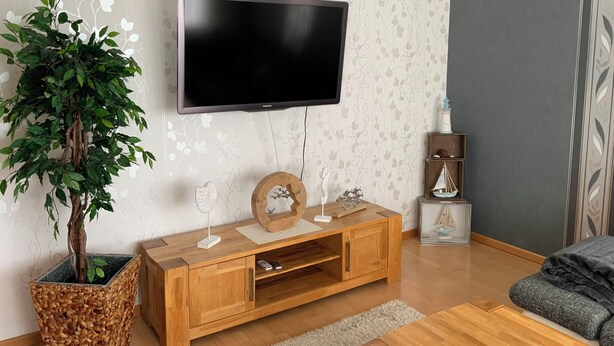 Un salon avec un téléviseur à écran plat fixé au mur, un meuble en bois orné d’objets décoratifs, une plante en pot et une petite étagère sur laquelle est posé un modèle de bateau.